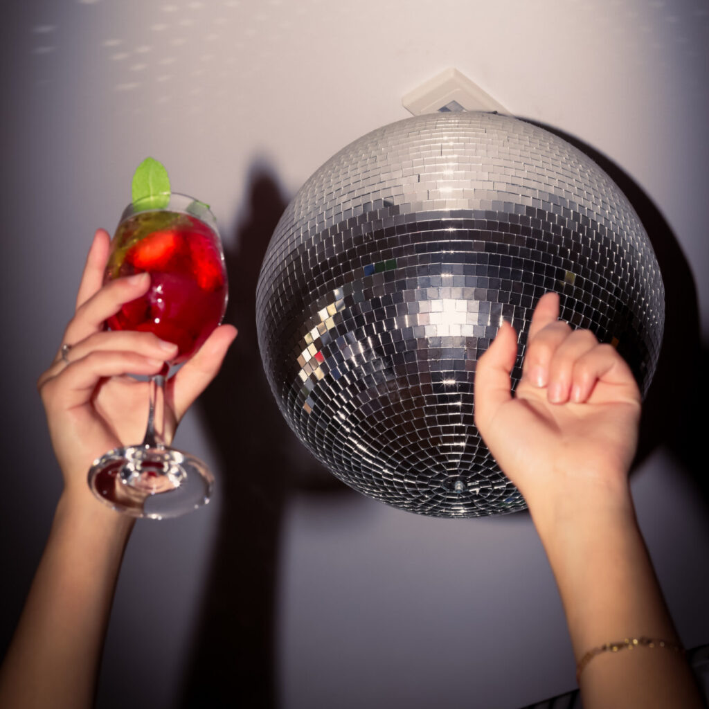 close up of hands pointed towards the ceiling in celebration. one hand is holding a cocktail beverage. there is a disco ball in the background