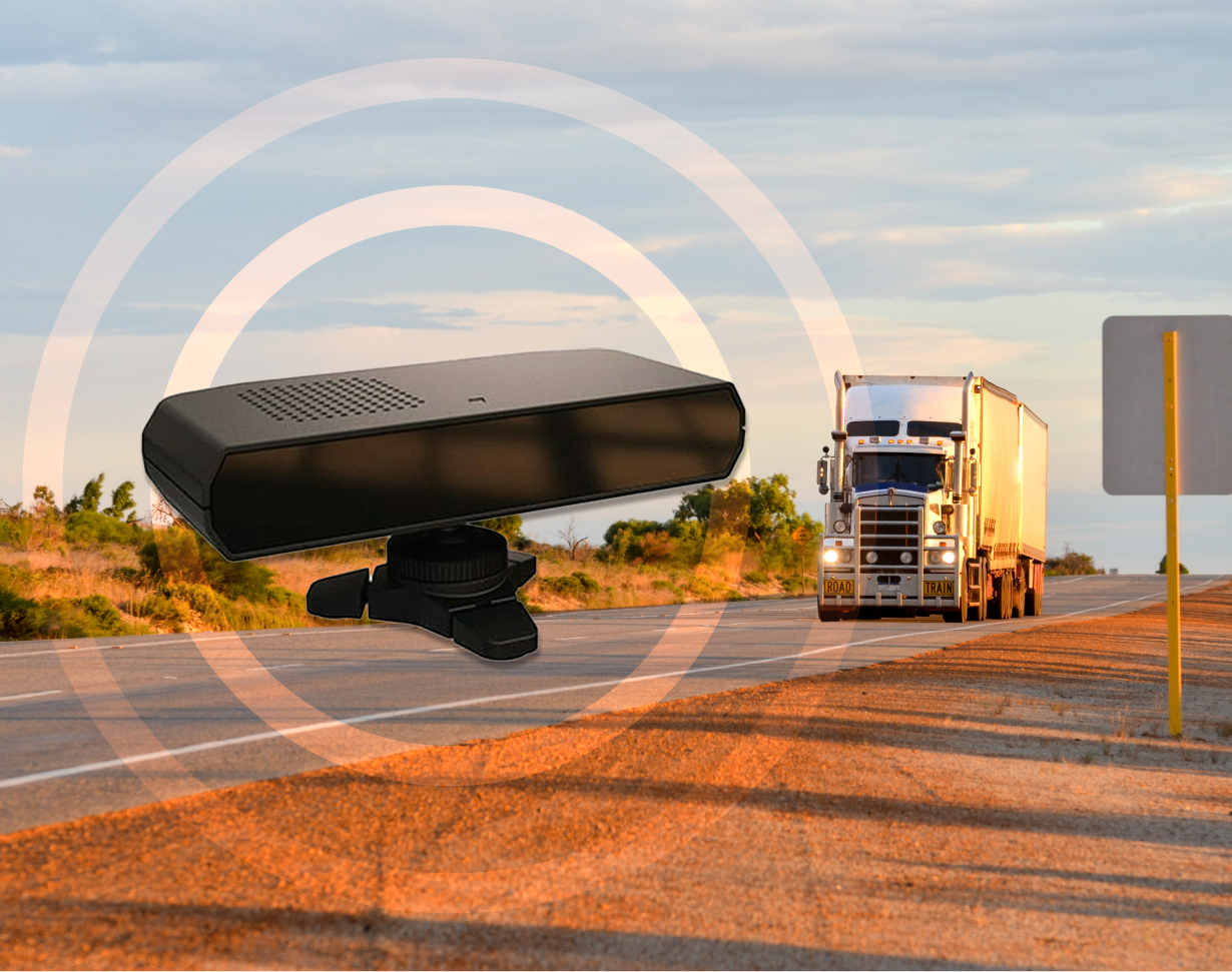 A semi-truck drives down a rural highway at sunset; in the foreground, a large black dashcam device is superimposed with concentric circles, suggesting surveillance or monitoring.